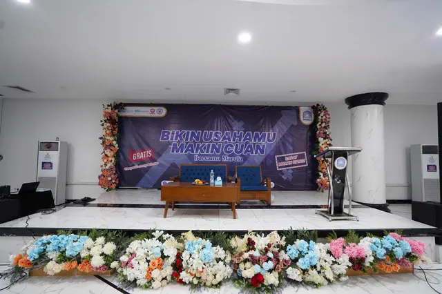 Panggung Aula Serbaguna ditata untuk seminar, dengan dekorasi bunga dan podium